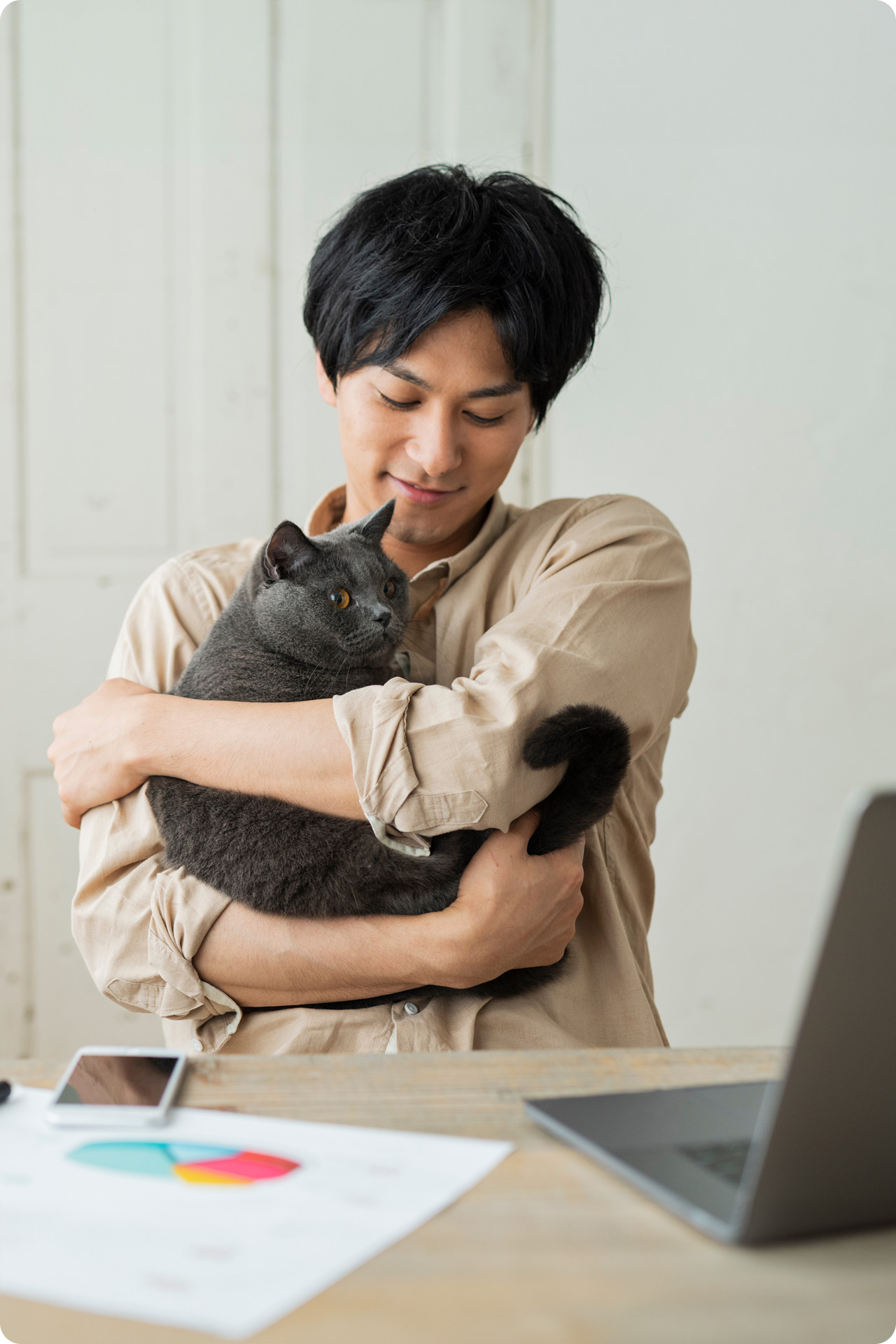 man holding a cat while sitting at work desk with laptop, paperwork, and phone